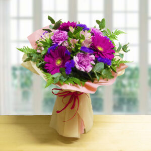 Bouquet Sweet Colors con Gerbere fucsia intenso, garofani viola e alstroemeria rosa, avvolto in carta regalo rosa e beige.