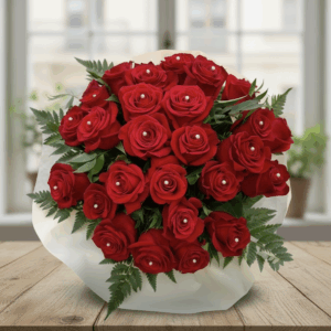 Bouquet di rose rosse "Pearl" in vaso di vetro, con dettagli perlati e confezione avorio, su tavolo in legno davanti a finestra sfocata.