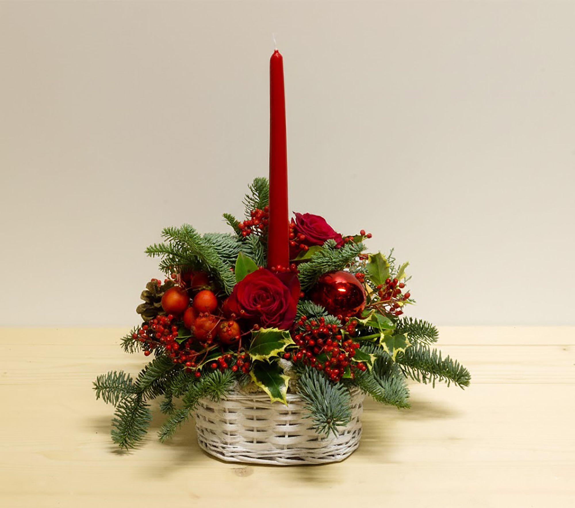 Centrotavola natalizio Happy Christmas con candela rossa, rose rosse, bacche, abete e pigne in cestino tondo bianco.