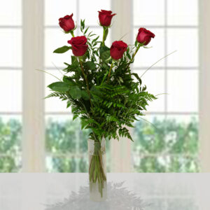 Bouquet di 5 rose rosse fresche con foglie di felce verde in un vaso di vetro cilindrico su fondo luminoso sfocato.
